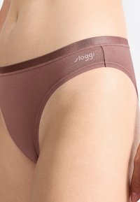 Close-up van een persoon die bruin low-rise ondergoed draagt met zichtbare naden en het "sloggi" logo op de tailleband.