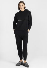 Czarna bluza z kapturem, wykonana z miękkiego materiału, z metaliczną paskiem na klatce piersiowej. W zestawie z czarnymi joggerami i szarymi butami wsuwanymi.