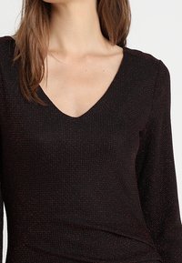 Černý top s výstřihem do V, dlouhými rukávy a jemnými červenými tečkami. Měkká textura a přiléhavý design zpevňují siluetu.