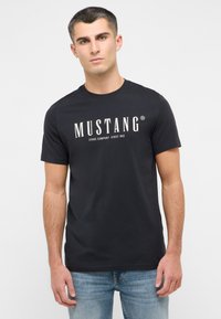 Melnas kokvilnas t-krekls ar baltu tekstu "MUSTANG" un saukli. Klasisks apaļais kakls, īsās piedurknes un mīksta auduma tekstūra.