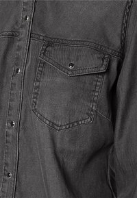 Camicia in denim nero con bottoni a pressione, tasca sul petto e dettagli di cucitura in vista. Il tessuto ha una finitura texturizzata.