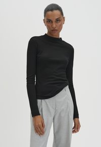 Frau mit zurückgebundenen Haaren, die ein eng anliegendes schwarzes langärmliges Oberteil und lockere hellgraue Hosen trägt, steht vor einem schlichten hellen Hintergrund.