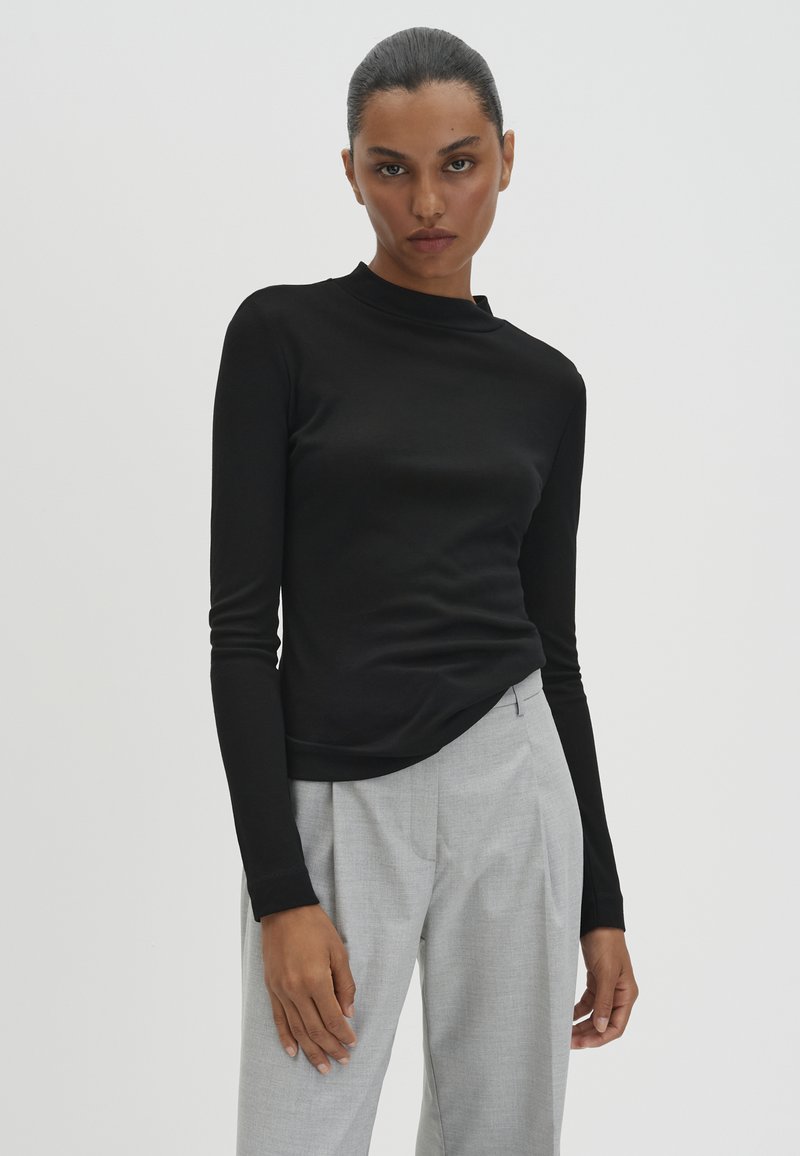 Frau mit zurückgebundenen Haaren, die ein eng anliegendes schwarzes langärmliges Oberteil und lockere hellgraue Hosen trägt, steht vor einem schlichten hellen Hintergrund.