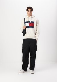 Cream hoodie with navy and red logo, paired with black cargo pants and white sneakers. Model holds a small black bag.