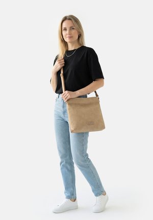 Vrouw staand, draagt een zwart shirt, lichtblauwe spijkerbroek, witte sneakers, houdt een beige crossbody-tas over haar schouder tegen een effen achtergrond.