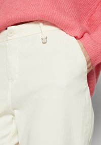 Λευκά βαμβακερά παντελόνια με λεία υφή και κουμπί κλεισίματος, συνδυασμένα με μια ανοιχτό ροζ πουλόβερ με υφή. Ο πάκος είναι ορατός.