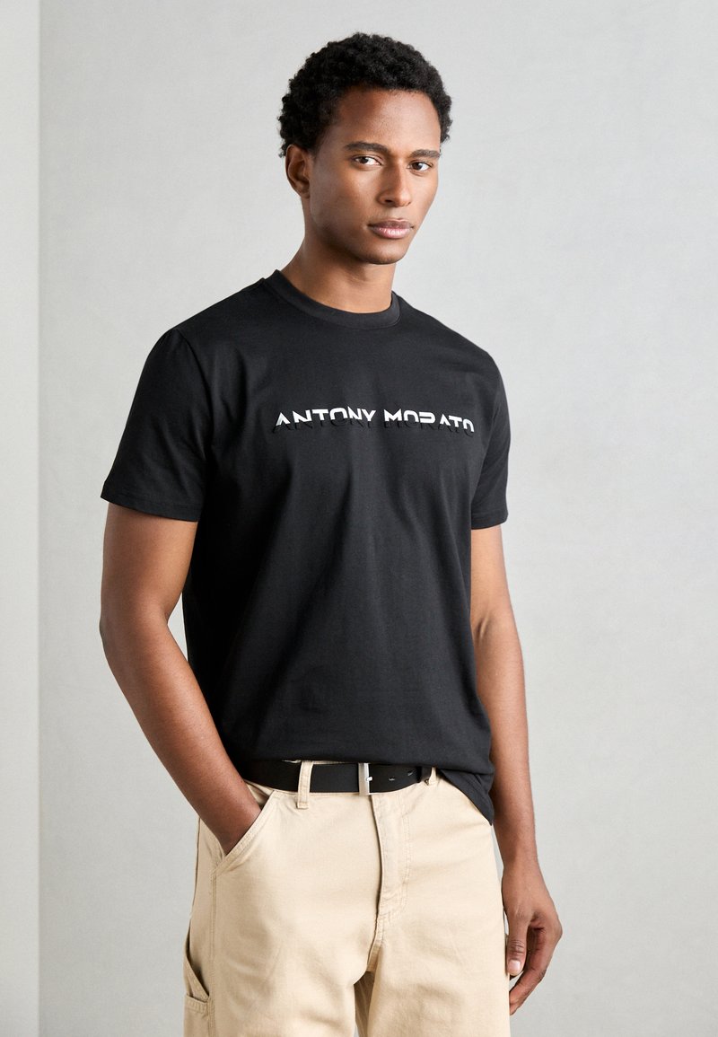 Zwarte katoenen t-shirt met het logo "ANTONY MORATO" in het wit, ronde hals, korte mouwen. Draagt met beige broek en zwarte riem. Glad van structuur.