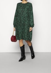 Vestido abstracto verde con mangas largas, cintura fruncida, combinado con botas negras hasta la rodilla y un bolso rojo con cadena dorada.