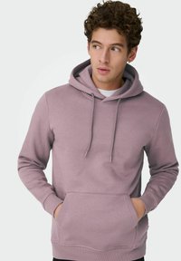 Jonge man met krullend haar, gekleed in een mauve hoodie met handen in de voorkantzak, kijkt opzij, tegen een effen lichte achtergrond.