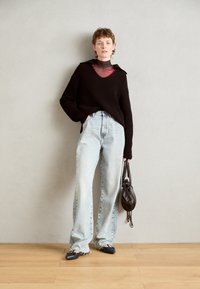 Suéter negro de punto acanalado con escote en V, jeans de tiro alto en azul claro y bailarinas negras. Sosteniendo un bolso de cuero oscuro. Suelo de madera.