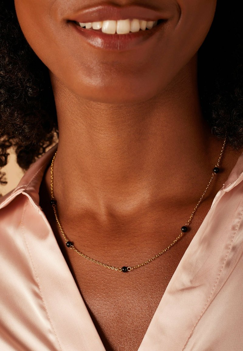 Collier en chaîne dorée avec des perles noires espacées régulièrement, porté sur un cou lisse. La chemise est en satin rose clair avec un col.