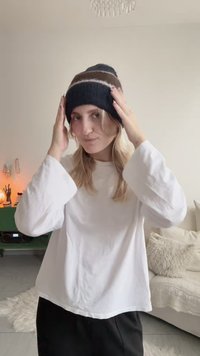 Schwarze und braune gestrickte Mütze mit horizontalen Streifen. Weißes Langarmshirt, strukturierter Hintergrund mit weichen Einrichtungsgegenständen.