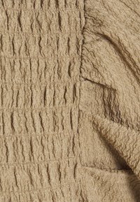 Beige stof met een gestructureerde, geplooide textuur en golvende stiknaden die een gekreukeld patroon over het oppervlak creëren.