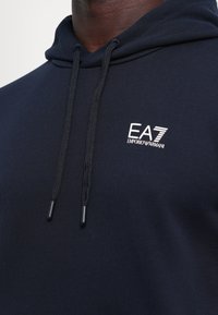 Felpa blu navy con cappuccio regolabile, tessuto morbido e logo ricamato EA7 Emporio Armani sul petto.