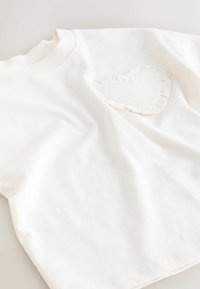 Weißes Baumwoll-T-Shirt mit einem strukturierten, herzförmigen Aufnäher mit einer gerafften Kante, der auf der linken Brustseite positioniert ist.