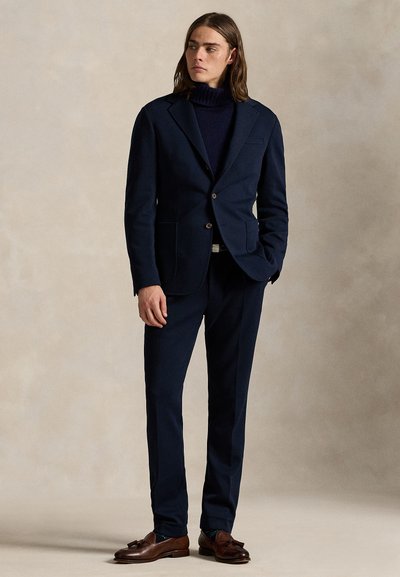 Man wearing a navy blue suit with a turtleneck sweater and brown tassel loafers standing against a plain neutral background.