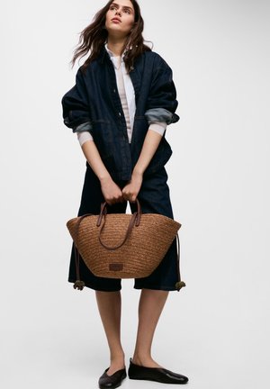 Kobieta ubrana w ciemną kurtkę dżinsową i spódnicę, trzyma dużą plecioną torbę na ramię ze skórzanymi rączkami, stoi na jednolitym tle.