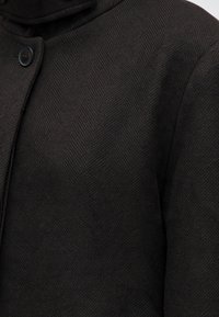 Gros plan d'un manteau en tissu chevrons noir avec un bouton noir près du col côté gauche.