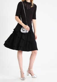 Svart topp med spetskrage, lager av svart kjol och silvriga klackskor med ankelrem. Inkluderar en liten silvrig crossbody-väska.