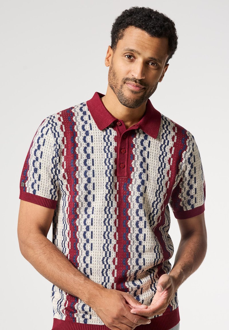 Polo de manga corta con cuello y puños en color burdeos, con un patrón geométrico en crema, azul marino y burdeos. La textura es tejida.