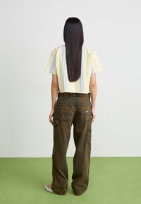 Camicia corta a righe gialle e bianche, abbinata a pantaloni cargo larghi marroni con tasche, vista da dietro su uno sfondo chiaro.