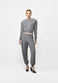 Naine seisab halli kudumisega s문의, seljas joggingupüksid ja mustad kõrged kontsad, lihtsa valge tausta taustal.