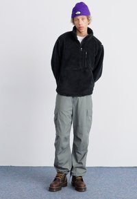 Pull en polaire noir avec une fermeture éclair quart, pantalon cargo gris et bottes lacées marron. Le modèle porte un bonnet en tricot violet. Fond neutre.