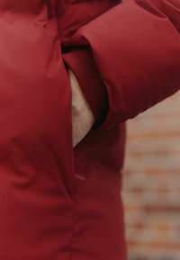 Červená nafukovacia bunda s širokým manžetou, so zreteľnou elastickou textúrou. Tkanina vyzerá hladko, s jemným leskom a minimálnym kovovým príslušenstvom.