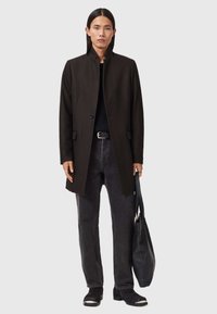 Musta villakangastakki korkean kauluksen kanssa, yksipainonappi suljettu, päällä teksturoitu musta neule ja tummat farkut, pidellen mustaa kassi.