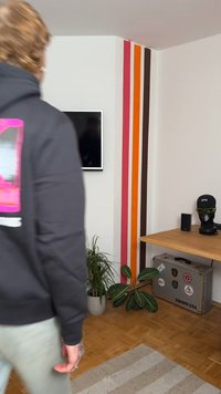 Crna dukserica s grafičkim dizajnom, svijetlozelene hlače, drveni stol, saksijska biljka i šarene zidne pruge u narančastoj, ružičastoj i smeđoj boji.