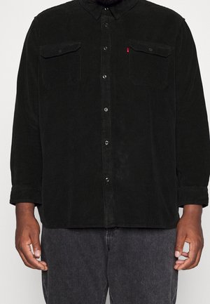 Homme portant une chemise noire en velours côtelé avec deux poches poitrine et un pantalon gris foncé, debout devant un fond uni.
