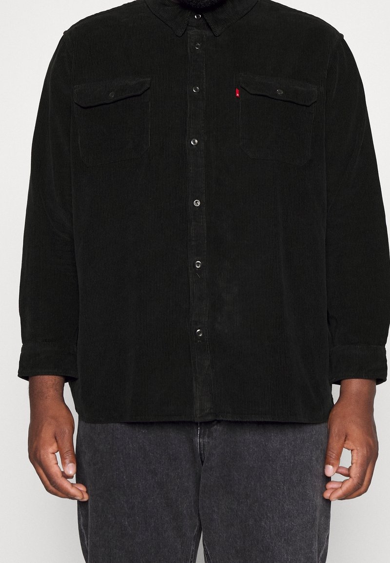 Homme portant une chemise noire en velours côtelé avec deux poches poitrine et un pantalon gris foncé, debout devant un fond uni.