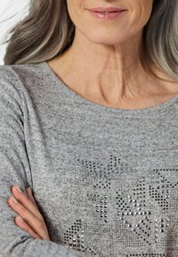 Grau gestrickter Pullover mit metallischen Nietenverzierungen in verschiedenen geometrischen Formen, rundem Ausschnitt und langen Ärmeln. Glatte Textur.
