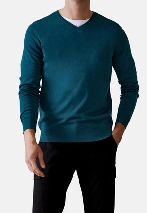Pull en v couleur bleu sarcelle en tissu doux, à manches longues et avec des poignets côtelés. Porté par-dessus une chemise blanche, associé à un pantalon noir.