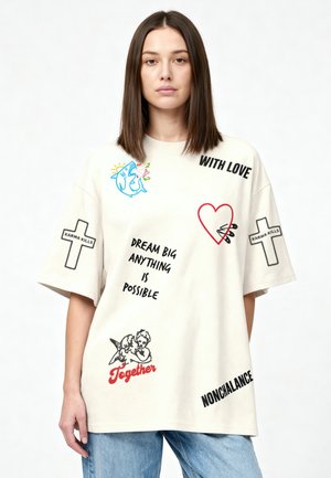 Frau trägt übergroßes weißes T-Shirt mit Hai, Herz, Engeln, Kreuzen und Text: „GROß TRÄUMEN, ALLES IST MÖGLICH“, „MIT LIEBE“, „NONCHALANCE“, „KARMA TÖTET“, „Zusammen.“