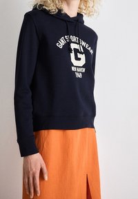 Personne portant un sweat à capuche bleu marine avec le texte « GANT SPORTSWEAR NEW HAVEN 1949 » et une jupe orange fendue à l'avant.