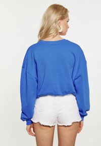 Sudadera azul de gran tamaño con hombros caídos, combinada con pantalones cortos de mezclilla blanca con bordes deshilachados y bolsillos traseros.