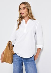 Chemise à col blanc avec patte de boutonnage, manches longues retroussées, assortie à un jean bleu et un sac en daim beige. Texture du tissu lisse.
