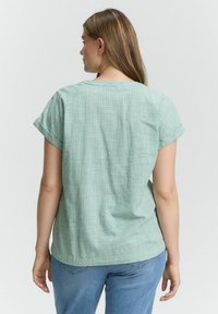 Light green, textured fabric t-shirt with rolled short sleeves and a relaxed fit, paired with blue denim jeans. Back view shown.