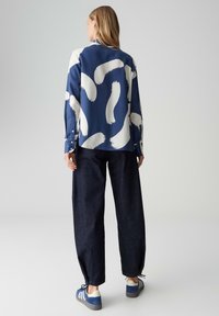 Langärmlige blaue Bluse mit weißem Pinselstrichmuster, kombiniert mit dunkelblauen, weiten Denim-Hosen und blauen Turnschuhen mit weißen Akzenten.