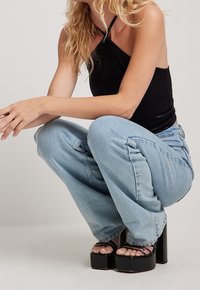 Top halter nero, jeans a gamba larga azzurri chiari e tacchi alti neri. Texture evidenti nei tessuti e design voluminoso delle scarpe.