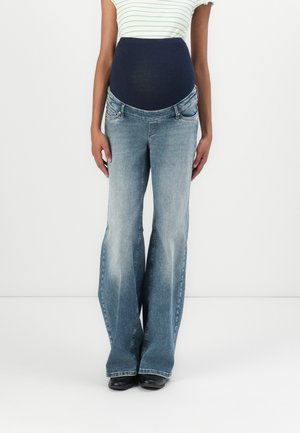Femme portant un jean de maternité évasé bleu clair avec une bande ventre bleu foncé et un haut blanc rayé, debout sur fond blanc.