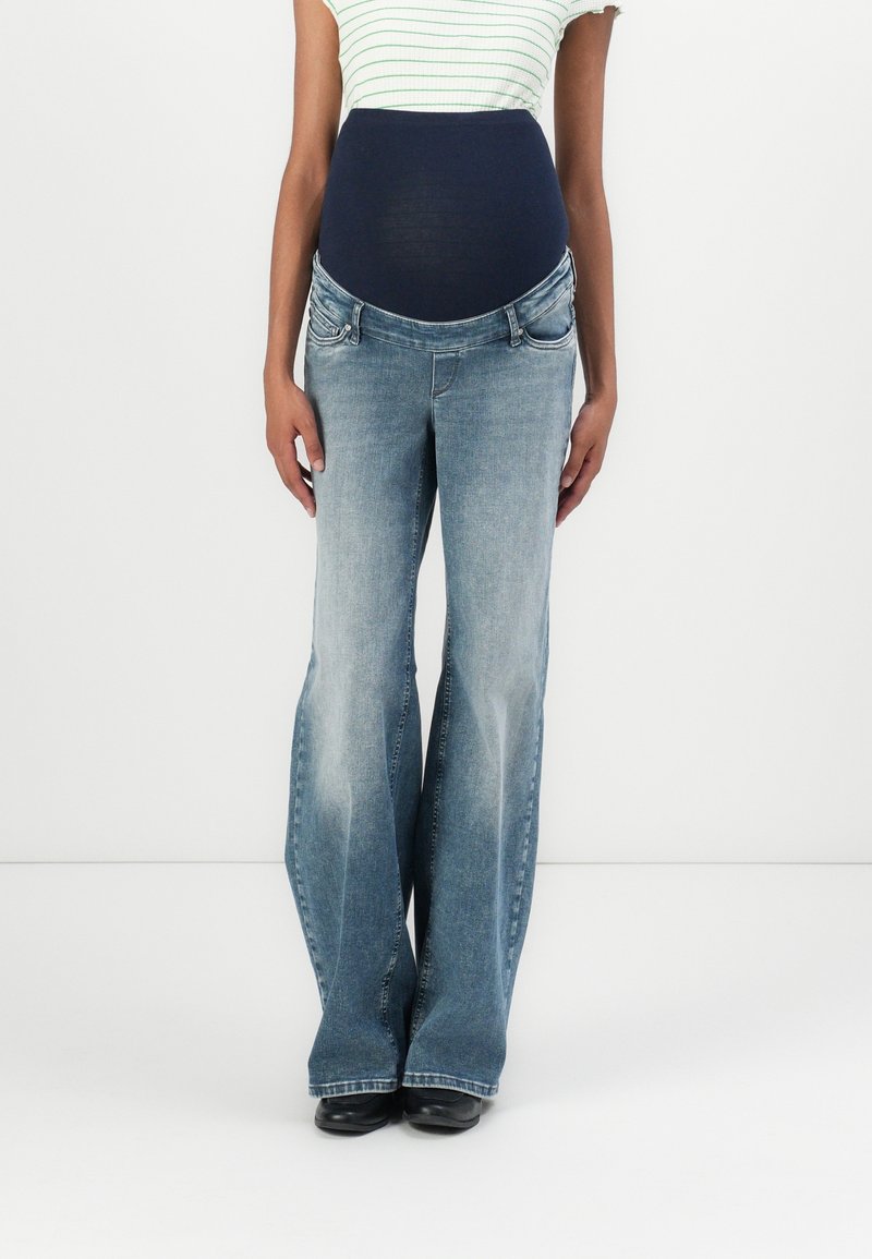 Femme portant un jean de maternité évasé bleu clair avec une bande ventre bleu foncé et un haut blanc rayé, debout sur fond blanc.