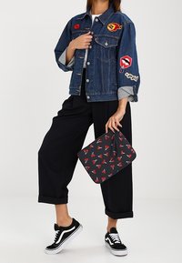 Veste en jean avec des patchs (flamme, lèvres) associée à un pantalon noir à jambes larges et des baskets noires ; tenant une pochette à motifs avec un dessin de lèvres rouges.
