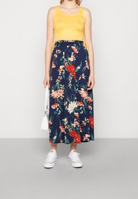 Gul ärmlös topp tillsammans med en marinblå blommig kjol med röda, vita och gröna mönster. Vita sneakers och en liten vit väska.