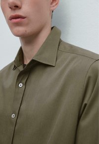 Camicia verde oliva con colletto, caratterizzata da una texture liscia e bottoni visibili. Il tessuto appare leggero e sagomato.