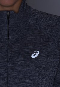 Donkergrijze zip-up jack van gestructureerde stof met een lichte heatherpatroon. Heeft een klein wit logo aan de bovenkant aan de linkerkant.