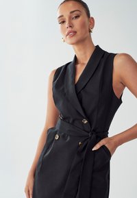 Abito nero senza maniche con colletto sciallato, dettagli a doppio petto e cintura legata in vita. Tessuto liscio e vestibilità sartoriale.
