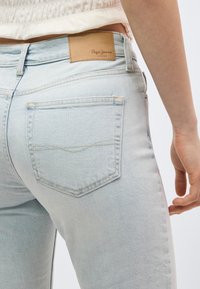 Helesinised teksapükste tagatasku lähivaade, millel on beež nahast Pepe Jeans Londoni silt ning osa inimese käest puusa kõrvalt.