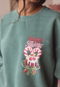 Žalias džemperis su šviesia tekstūra, vaizduojantis raudonai baltą saldainio lazdelės dizainą, tortą su glaistu ir tekstą „SEASONS SWEET THINGS“.